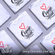 Футболка Серце знає, біла, зображена на моделі в ракурсі зліва, фон світло-фіолетовий, принт з червоним серцем і чорним написом 'Серце знає'.