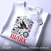 Футболка Шлях життя. Біла футболка, складена на поверхні, з видимим принтом, що містить птахів, квіти та сонце. На футболці є етикетка з зеленим шнурком, фон світлий і однорідний.