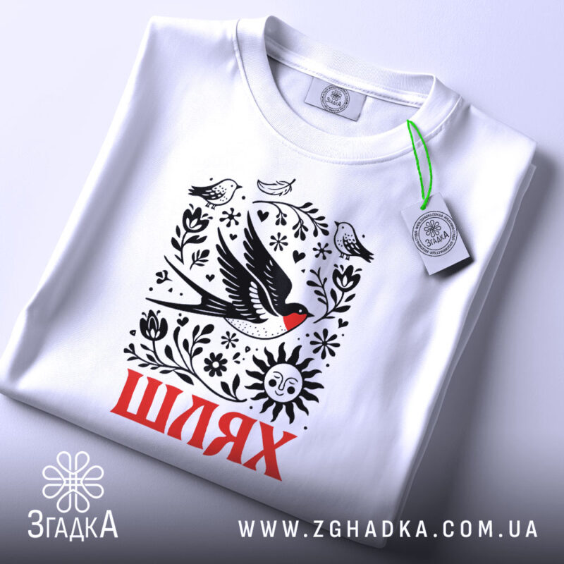 Футболка Шлях життя. Біла футболка, складена на поверхні, з видимим принтом, що містить птахів, квіти та сонце. На футболці є етикетка з зеленим шнурком, фон світлий і однорідний.