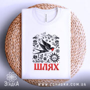 Футболка Шлях життя. Біла футболка, розкладена на плетеній кошику, з яскравим принтом, що включає птахів, сонце та слово 'Шлях' червоним кольором. Фон світлий, що створює контраст з текстурою кошика.