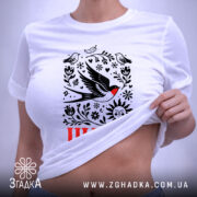 Футболка Шлях життя. Біла футболка, складена на поверхні, з видимим принтом, що містить птахів, квіти та сонце. На футболці є етикетка з зеленим шнурком, фон світлий і однорідний.