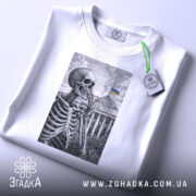 Футболка Скелет і Верховна Рада, біла футболка на столі, зображення скелета на фоні будівлі Верховної Ради з українським прапором, принт виконаний у чорному кольорі, на футболці є етикетка з логотипом бренду, фон світлий.