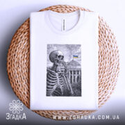 Футболка Скелет і Верховна Рада, біла футболка на плетеній кошику, зображення скелета на фоні будівлі Верховної Ради з українським прапором, принт виконаний у чорному кольорі, фон світлий.