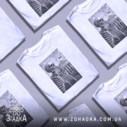 Футболка Скелет і Верховна Рада, біла футболка на моделі, зображення скелета на фоні будівлі Верховної Ради з українським прапором, принт виконаний у чорному кольорі, модель позує з легким усмішкою, фон світло-фіолетовий.