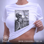 Футболка Скелет і Верховна Рада, біла футболка на столі, зображення скелета на фоні будівлі Верховної Ради з українським прапором, принт виконаний у чорному кольорі, на футболці є етикетка з логотипом бренду, фон світлий.