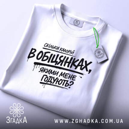 Футболка скільки калорій в обіцянках, біла футболка, що лежить на поверхні, фон світло-сірий, принт чорного кольору з текстом, що запитує про кількість калорій в обіцянках, з етикеткою зеленого кольору.