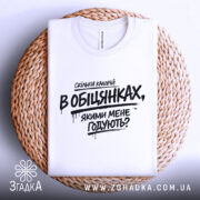 Футболка скільки калорій в обіцянках, біла футболка, що лежить на плетеній основі, фон світло-сірий, принт чорного кольору з текстом, що запитує про кількість калорій в обіцянках.