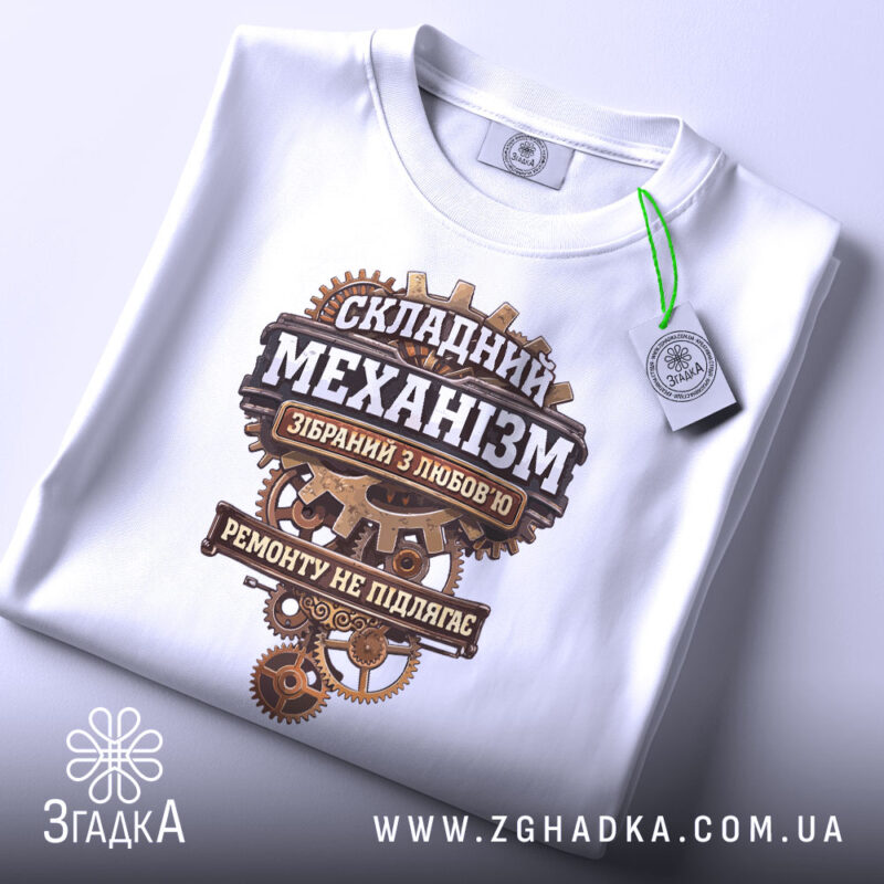 Футболка Складний механізм, біла, на плоскій поверхні, ракурс зверху, фон світло-фіолетовий, принт з механічними деталями та текстом, що говорить про складність механізму, з етикеткою.