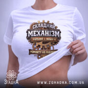 Футболка Складний механізм, біла, на плоскій поверхні, ракурс зверху, фон світло-фіолетовий, принт з механічними деталями та текстом, що говорить про складність механізму, з етикеткою.