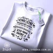 Футболка сміх і секс продовжують життя, біла, зображена зверху, ракурс зверху, фон світло-фіолетовий, принт чорного кольору з текстом і декоративними елементами, на футболці є етикетка.