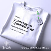 Футболка Спочатку ти прочитаєш, біла, на столі, з етикеткою, фон світлий, принт чорного кольору з текстом, розташованим по центру.