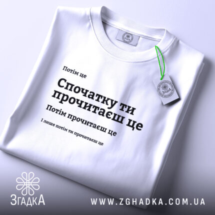 Футболка Спочатку ти прочитаєш, біла, на столі, з етикеткою, фон світлий, принт чорного кольору з текстом, розташованим по центру.