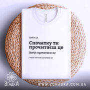 Футболка Спочатку ти прочитаєш, біла, складена на плетеній кошику, фон світлий, принт чорного кольору з текстом, розташованим по центру.