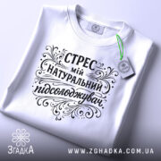 Футболка Стрес мій підсолоджувач, біла, лежить на столі, з верхнього ракурсу, фон світло-сірий, принт чорного кольору з витонченими декоративними елементами, на комірці є етикетка.