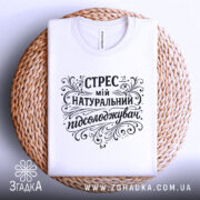Футболка Стрес мій підсолоджувач, біла, лежить на плетеній основі, з верхнього ракурсу, фон світло-сірий, принт чорного кольору з витонченими декоративними елементами, на комірці є етикетка.