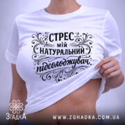 Футболка Стрес мій підсолоджувач, біла, лежить на столі, з верхнього ракурсу, фон світло-сірий, принт чорного кольору з витонченими декоративними елементами, на комірці є етикетка.