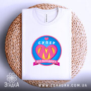 Футболка супер мама, біла, складена на плоскій поверхні, фон світло-сірий, принт з написом 'СУПЕР' та серцем, кольори принта: рожевий, жовтий, блакитний, на плоскій кошику.