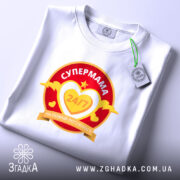 Футболка супермама білого кольору, розкладена на поверхні. Принт яскравий, з червоним та жовтим кольорами, з текстом 'СУПЕРМАМА 24/7' у серці. На футболці видно етикетку з логотипом бренду, фон світлий, що підкреслює деталі принта.