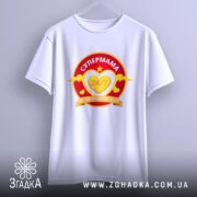 Футболка супермама білого кольору, підвішена на вішалці. Принт яскравий, з червоним та жовтим кольорами, містить текст 'СУПЕРМАМА 24/7' у серці. Фон світлий, що створює чистий і свіжий вигляд.