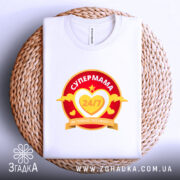 Футболка супермама білого кольору, складена на плетеній кошику. Принт яскравий, з червоним та жовтим кольорами, з текстом 'СУПЕРМАМА 24/7' у серці. Фон нейтральний, що підкреслює текстуру кошика.