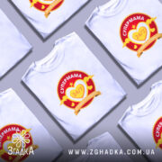 Футболка супермама білого кольору, зображена на моделі, яка позує з легким усмішкою. Принт на футболці яскравий, з червоним та жовтим кольорами, містить текст 'СУПЕРМАМА 24/7' у серці. Фон м'який, пастельний, що підкреслює стиль футболки.