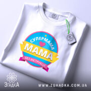 Футболка супермама без інструкції, біла, на плоскій поверхні, фон світло-фіолетовий, принт кольоровий з написом 'СУПЕРМАМА' та 'Мама без інструкцій', з етикеткою на комірці.
