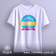 Футболка супермама без інструкції, біла, підвішена на вішалці, фон світло-сірий, принт кольоровий з написом 'СУПЕРМАМА' та 'Мама без інструкцій'.