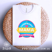 Футболка супермама без інструкції, біла, складена на круглому плетеному кошику, фон світло-фіолетовий, принт кольоровий з написом 'СУПЕРМАМА' та 'Мама без інструкцій'.