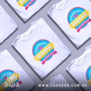 Футболка супермама без інструкції, біла, на моделі, з легким макіяжем та укладкою, фон світло-фіолетовий, принт кольоровий з написом 'СУПЕРМАМА' та 'Мама без інструкцій'.