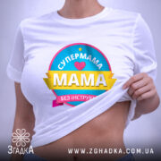 Футболка супермама без інструкції, біла, на плоскій поверхні, фон світло-фіолетовий, принт кольоровий з написом 'СУПЕРМАМА' та 'Мама без інструкцій', з етикеткою на комірці.