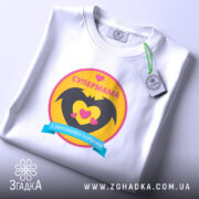 Футболка супермама, біла, на столі, ракурс зверху, фон світло-сірий, принт з жовтим колом, чорними крилами та сердечками, напис 'СУПЕРМАМА' у верхній частині, з етикеткою.