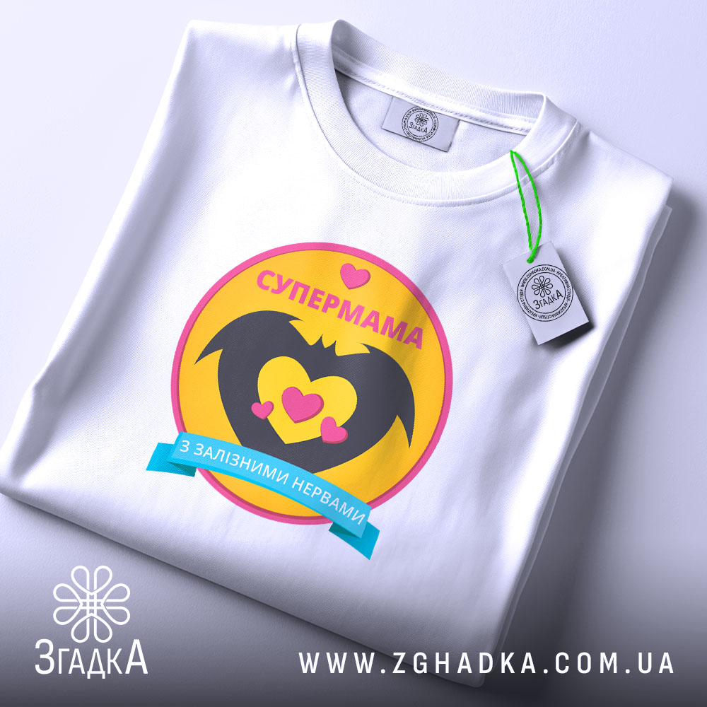 Футболка супермама, біла, на столі, ракурс зверху, фон світло-сірий, принт з жовтим колом, чорними крилами та сердечками, напис 'СУПЕРМАМА' у верхній частині, з етикеткою.