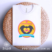 Футболка супермама, біла, на столі, ракурс зверху, фон світло-сірий, принт з жовтим колом, чорними крилами та сердечками, напис 'СУПЕРМАМА' у верхній частині, на плетеній основі.