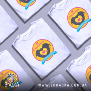 Футболка супермама, біла, на моделі, ракурс спереду, фон світло-фіолетовий, принт з жовтим колом, чорними крилами та сердечками, напис 'СУПЕРМАМА' у верхній частині.