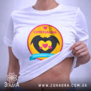 Футболка супермама, біла, на столі, ракурс зверху, фон світло-сірий, принт з жовтим колом, чорними крилами та сердечками, напис 'СУПЕРМАМА' у верхній частині, з етикеткою.