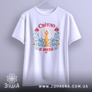 Футболка Світло в мені, біла, підвішена на вішалці, з принтом свічки та квітів, фон світло-сірий, деталі принта яскраві, з червоними, жовтими та зеленими елементами.