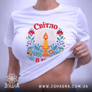 Футболка Світло в мені, біла, на столі, з принтом свічки та квітів, фон світло-сірий, деталі принта яскраві, з червоними, жовтими та зеленими елементами, з етикеткою.