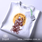 Футболка свобода дорожча, складена біла футболка з принтом, вид зверху, фон світло-сірий, принт з воїном та птахом, етикетка з зеленим шнурком.