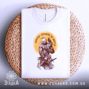 Футболка свобода дорожча, складена біла футболка на плетеній основі, вид зверху, фон світло-сірий, принт з воїном та птахом, кольори принта коричневі та жовті.