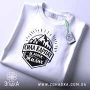 Футболка Сила Карпат, біла, на плоскій поверхні, з верхнього ракурсу, фон світло-фіолетовий, принт чорного кольору з горами та написом 'Сила Карпат у моїх жилах', з етикеткою зеленого кольору.