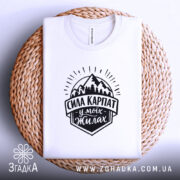 Футболка Сила Карпат, біла, на плоскій поверхні, з верхнього ракурсу, фон світло-фіолетовий, принт чорного кольору з горами та написом 'Сила Карпат у моїх жилах', розташована на плетеній основі.