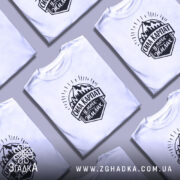 Футболка Сила Карпат, біла, на моделі, з легким ракурсом спереду, фон світло-фіолетовий, принт чорного кольору з горами та написом 'Сила Карпат у моїх жилах'.