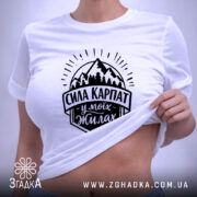 Футболка Сила Карпат, біла, на плоскій поверхні, з верхнього ракурсу, фон світло-фіолетовий, принт чорного кольору з горами та написом 'Сила Карпат у моїх жилах', з етикеткою зеленого кольору.