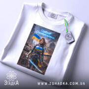 Футболка сильна українська жінка, біла, розкладена на поверхні, з легким ракурсом зверху, фон світлий, принт з героїнею в броні з мечем, на фоні зруйнованого міста та українським прапором, з етикеткою на комірці.