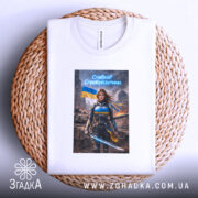 Футболка сильна українська жінка, біла, розкладена на плетеній основі, з легким ракурсом зверху, фон світлий, принт з героїнею в броні з мечем, на фоні зруйнованого міста та українським прапором.