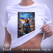 Футболка сильна українська жінка, біла, розкладена на поверхні, з легким ракурсом зверху, фон світлий, принт з героїнею в броні з мечем, на фоні зруйнованого міста та українським прапором, з етикеткою на комірці.