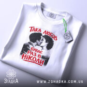 Футболка така любов, біла, на плоскій поверхні, з принтом, що зображує пару, яка цілується, текст червоним кольором: 'ТАКА ЛЮБОВ БУВАЄ РАЗ В НІКОЛИ', етикетка з зеленим шнурком, фон світлий і мінімалістичний.