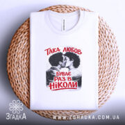 Футболка така любов, біла, на плоскій поверхні, з принтом, що зображує пару, яка цілується, текст червоним кольором: 'ТАКА ЛЮБОВ БУВАЄ РАЗ В НІКОЛИ', розташована на плетеній основі, фон світлий і природний.