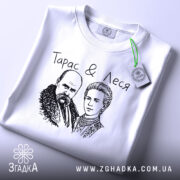 Футболка Тарас і Леся, біла, складена на поверхні, ракурс зверху, фон світло-сірий, принт з малюнком Тараса Шевченка та Лесі Українки, текст "Тарас & Леся" над малюнком, на етикетці зелена стрічка.