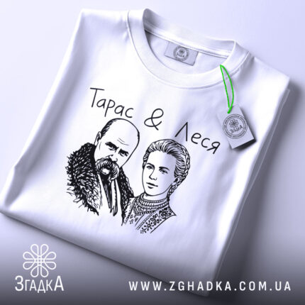 Футболка Тарас і Леся, біла, складена на поверхні, ракурс зверху, фон світло-сірий, принт з малюнком Тараса Шевченка та Лесі Українки, текст "Тарас & Леся" над малюнком, на етикетці зелена стрічка.
